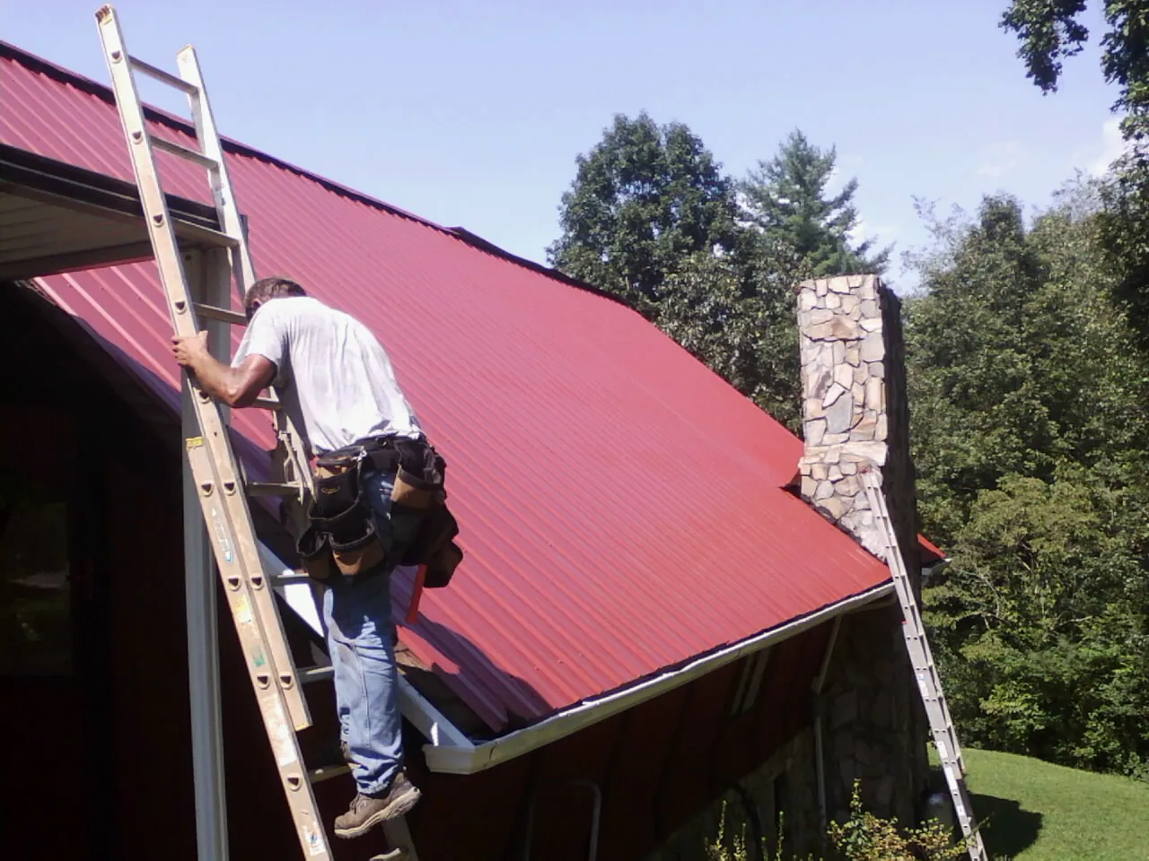 Roofer on a ladder beside a red metal roof and stone chimney — Soffit & Fascia Repair by Roofing Brothers in South Bend